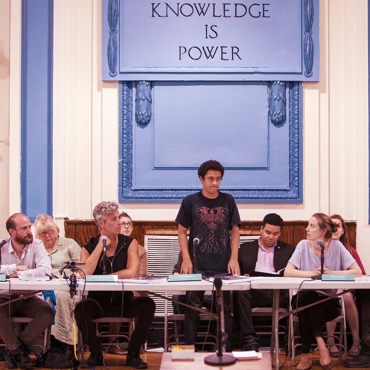 Man standing at a table of seated listeners beneath a blue sign that says knowledge is power